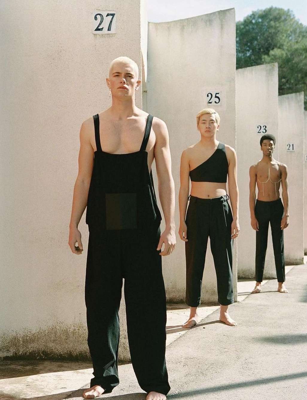 Three diverse models in fashionable black attire pose against a textured wall with numbered signs, creating a minimalist yet striking composition.