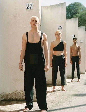 Three diverse models in fashionable black attire pose against a textured wall with numbered signs, creating a minimalist yet striking composition.