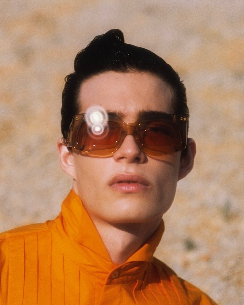 Close-up portrait of a young adult man wearing orange shirt and brown tinted sunglasses, set against a neutral outdoor background.