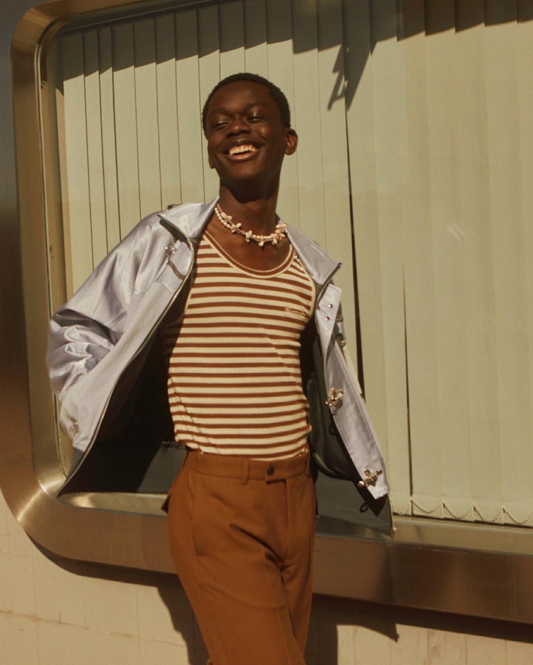 Smiling Black man in fashionable brown striped top, rust-colored pants, and blue jacket poses outdoors in front of window blinds.