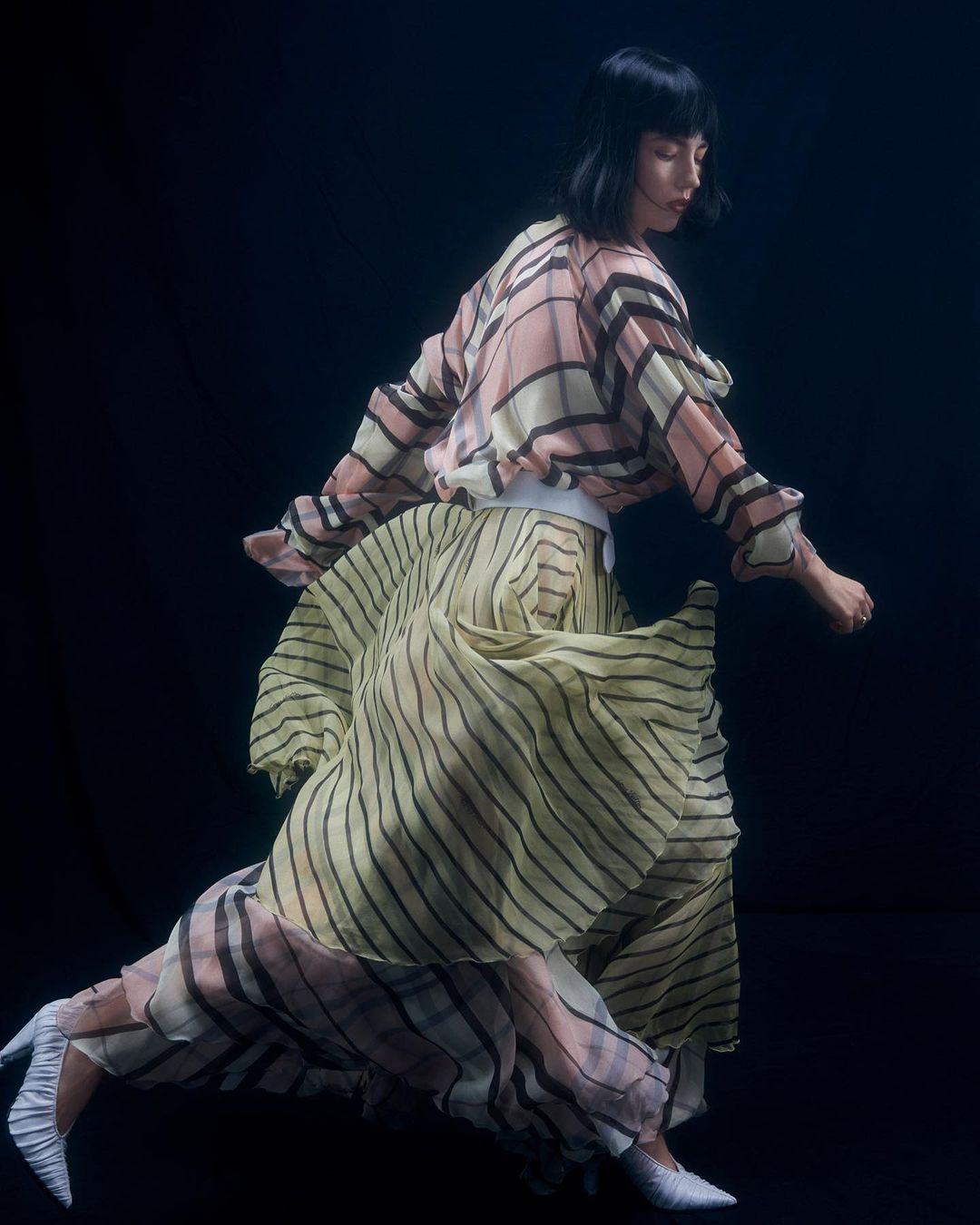 Woman in a patterned pleated dress and white heels moving against a dark background
