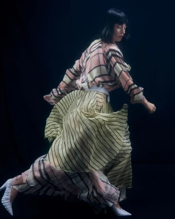 Woman in a patterned pleated dress and white heels moving against a dark background