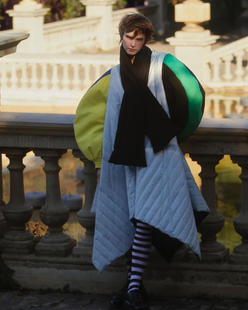 A person poses outdoors on a stone terrace wearing a large coat with yellow and green accents, a black scarf, and black-and-white striped tights.