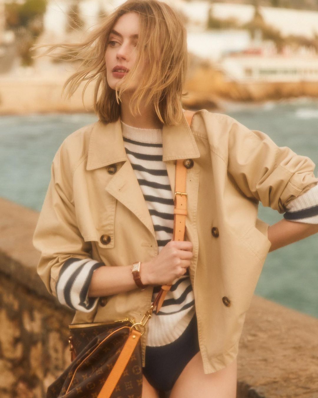 A woman wearing a tan trench coat and striped sweater stands outdoors near the coast, photographed by Sergi Pons.