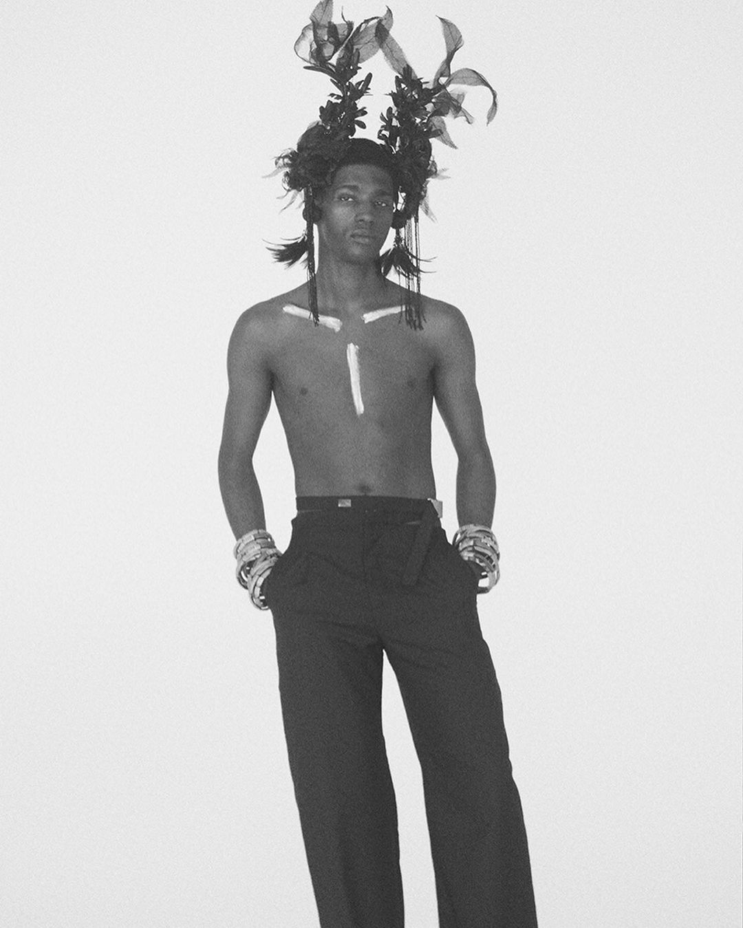 Monochrome studio shot of a slender, shirtless male model with headpiece, wristbands, and pants, posing in front of a light background.