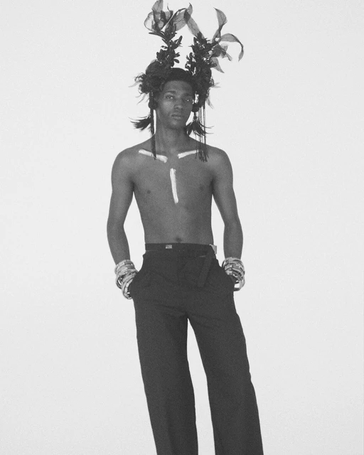 Monochrome studio shot of a slender, shirtless male model with headpiece, wristbands, and pants, posing in front of a light background.