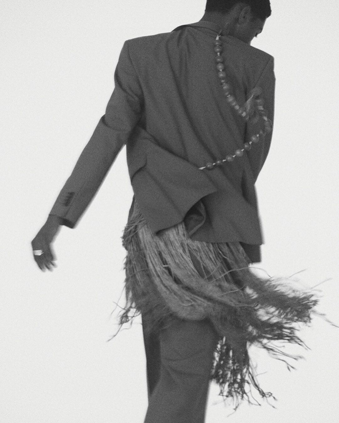 Back view of stylish adult male in a grey suit with fringe detail against a plain white background in a fashion-forward studio shot.