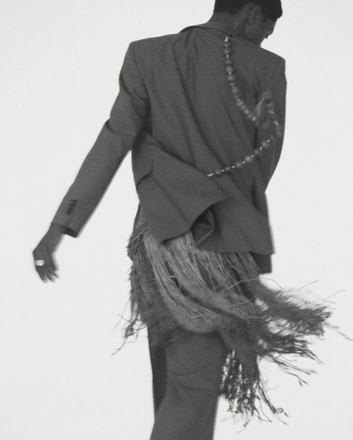 Back view of stylish adult male in a grey suit with fringe detail against a plain white background in a fashion-forward studio shot.