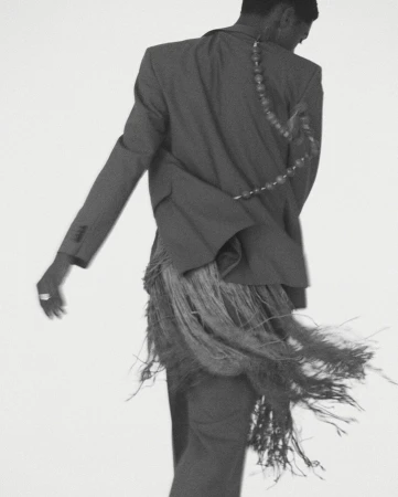 Back view of stylish adult male in a grey suit with fringe detail against a plain white background in a fashion-forward studio shot.