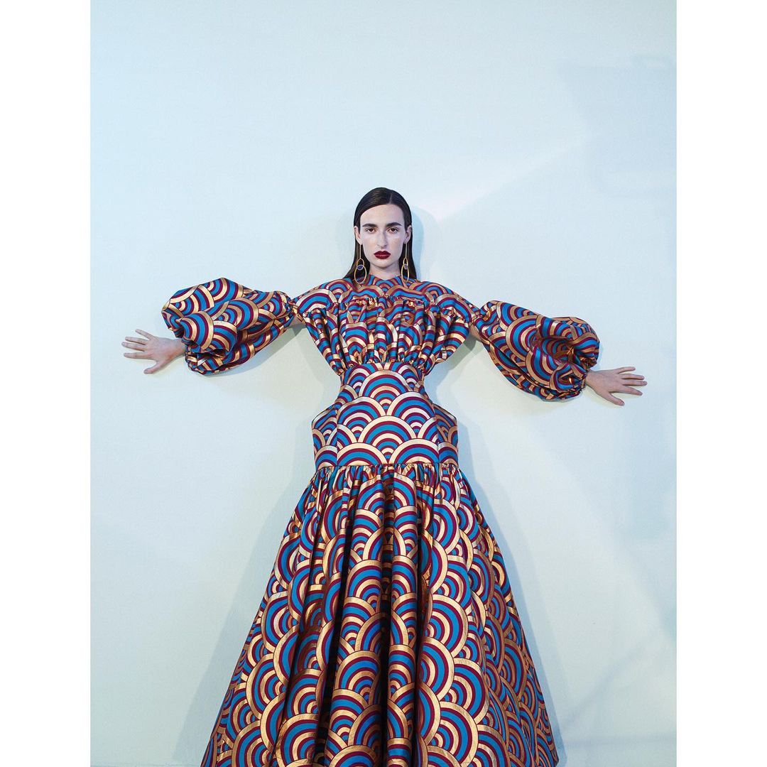 Adult female fashion model poses in a long blue and copper pattern dress with large sleeves against a light-colored backdrop.