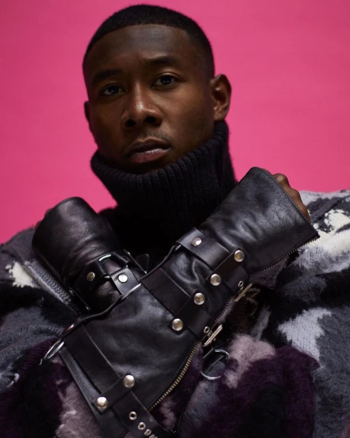Close-up shot of an adult man wearing a black turtleneck and leather wristbands against a vibrant pink backdrop, fashion portrait.