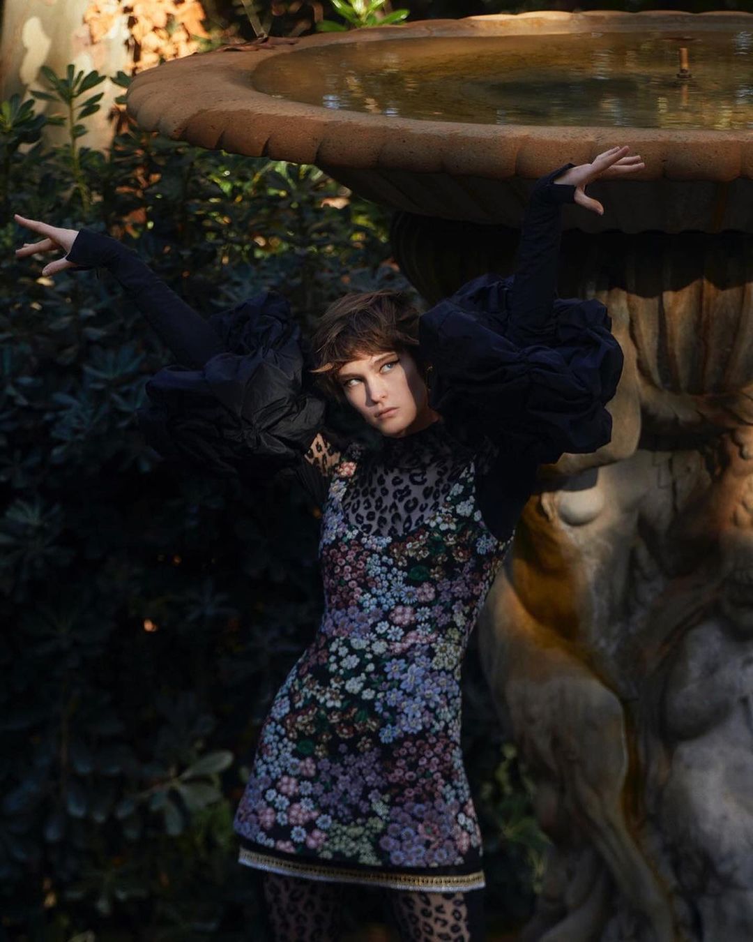 Woman in floral dress poses near stone fountain in sunlit garden. Fashion editorial image, with lush foliage in the background.