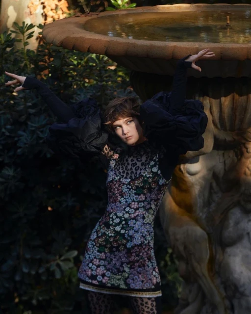 Woman in floral dress poses near stone fountain in sunlit garden. Fashion editorial image, with lush foliage in the background.