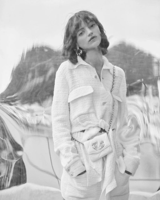 Woman in a white tweed jacket and skirt with a small chain bag poses in front of a blurry, reflective background.