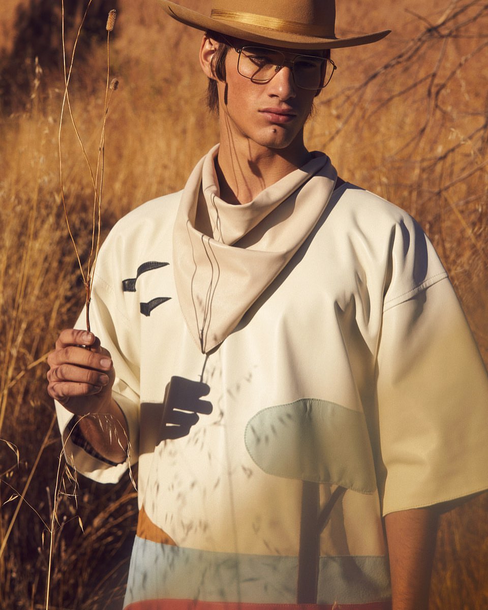 A man wearing a hat and oversized shirt poses in a dry field, holding grass stalks. He is wearing glasses and a cream-colored bandana.