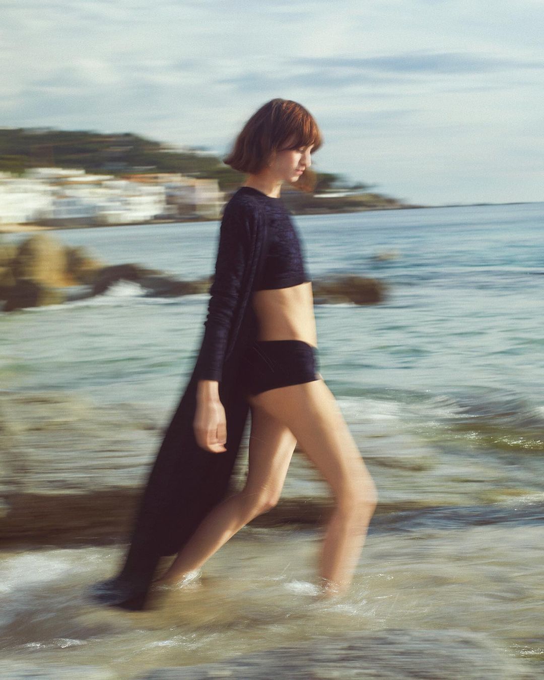 Woman in swimwear and cover-up walking on a beach with a blurred motion effect, showcasing fashion and a coastal landscape with rocky outcrops.
