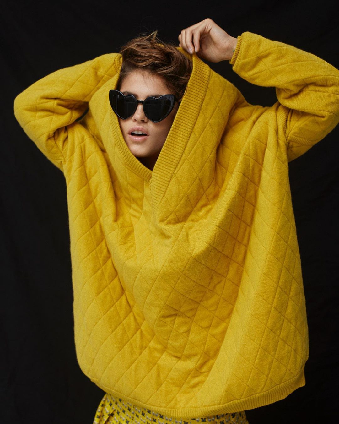 Stylish woman in a yellow oversized quilted sweater and heart-shaped sunglasses poses against a dark background, capturing a trendy fashion moment.
