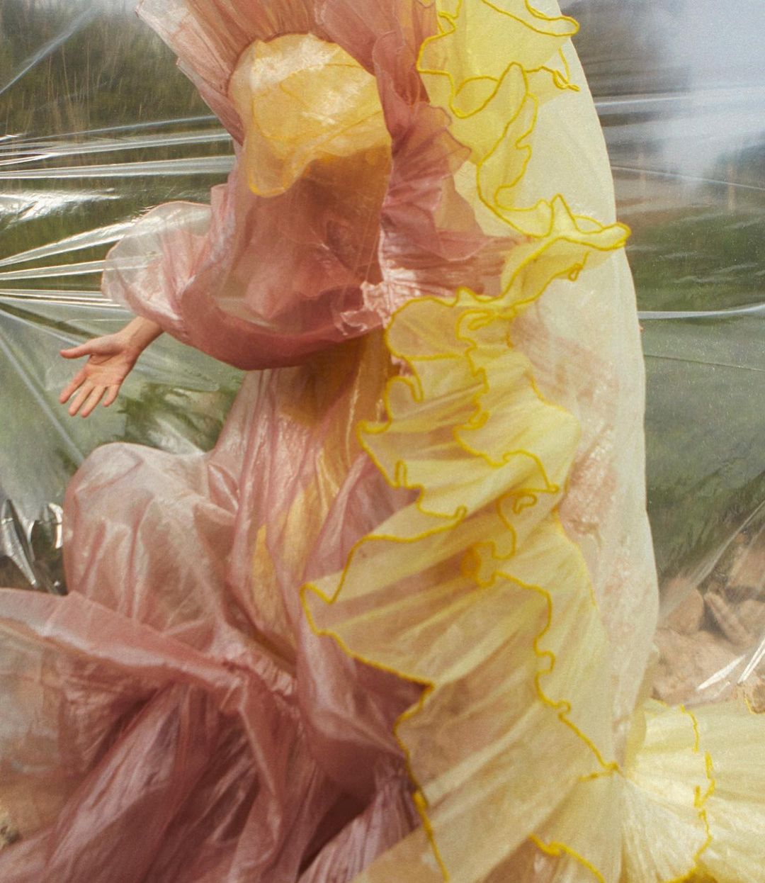 Model wearing a voluminous pink and yellow ruffled dress behind a layered transparent plastic sheet