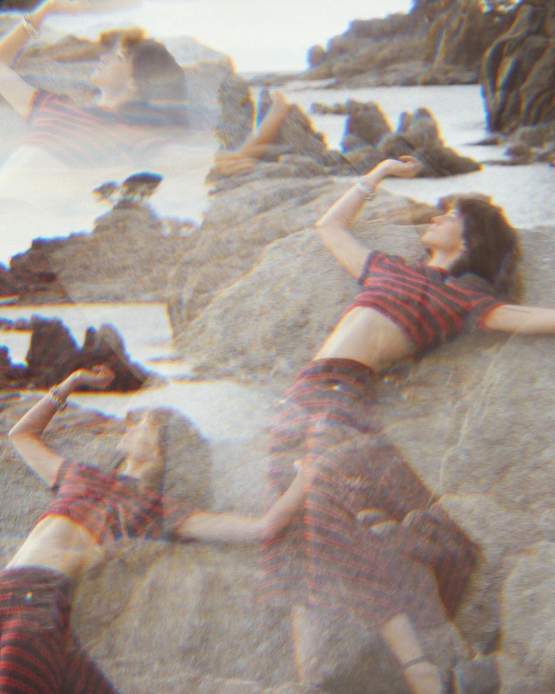 A woman in a striped outfit is lying on rocks near the ocean, with a multiple exposure effect creating a dreamy, ethereal vibe.