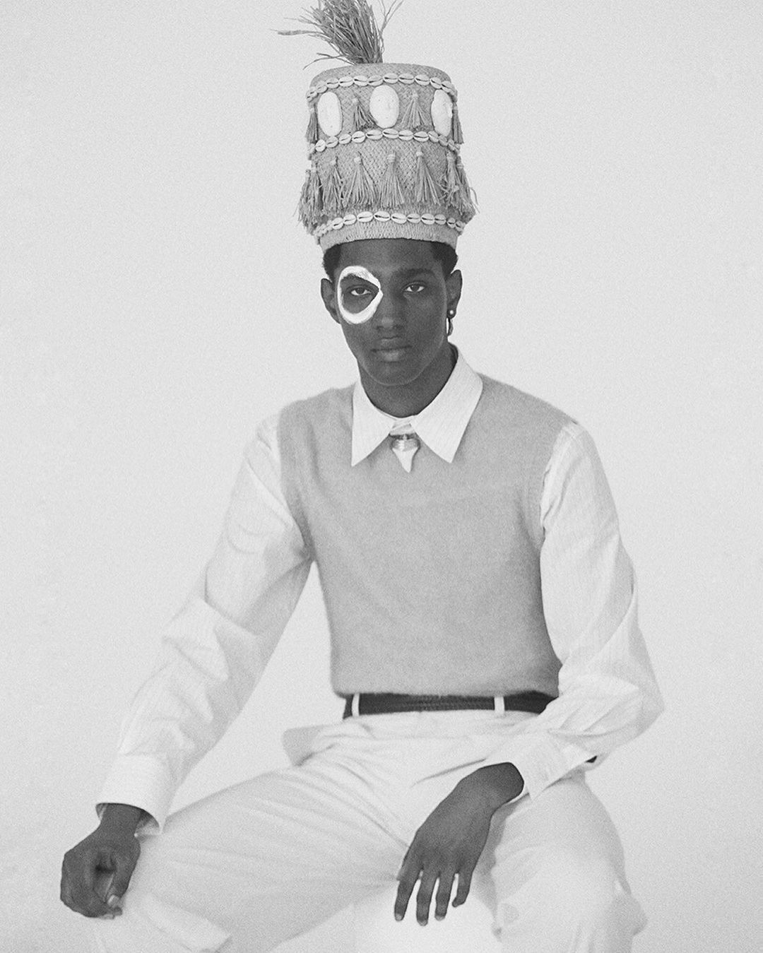 Black male model in a white shirt, vest, pants, and traditional straw hat with shells, posing in a photo studio with white backdrop.