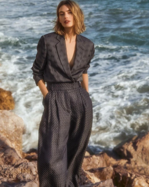 Adult woman poses in dark patterned suit on rocky coast with ocean backdrop, medium shot, natural light, windblown bob hairstyle.