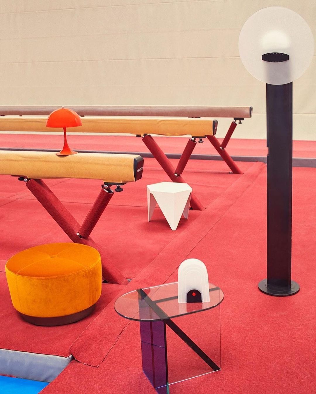Interior design featuring a balance beam, tables, a lamp, and seating arranged on a red floor in a brightly lit indoor space.