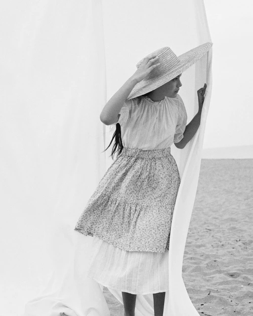 Young woman in a dress and straw hat poses with white fabric on a beach. Black and white portrait showcases fashion and style.