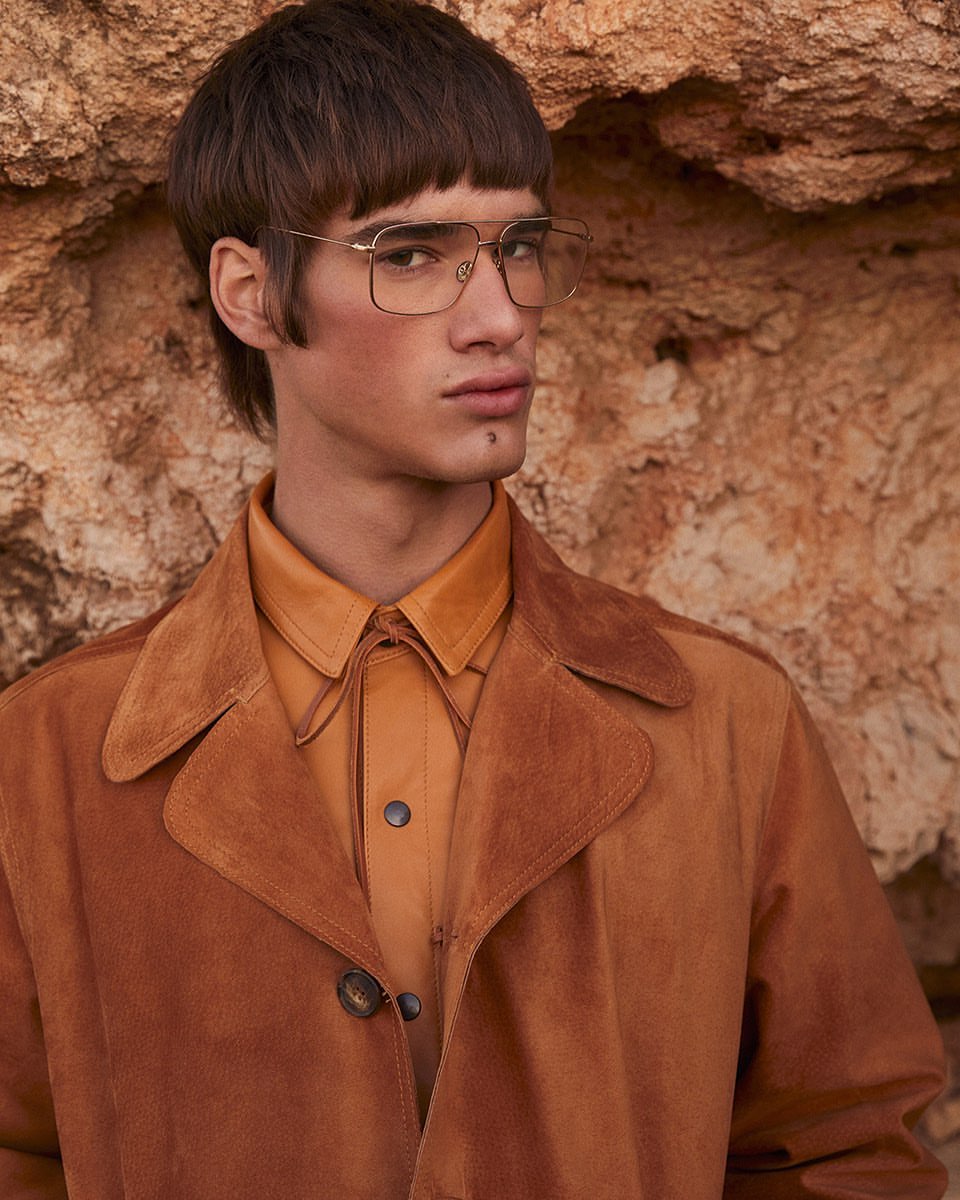 A stylish adult male with unique bowl-cut hairstyle, posing with glasses and brown suede coat against a textured, earthy-toned rock wall.