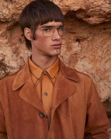 A stylish adult male with unique bowl-cut hairstyle, posing with glasses and brown suede coat against a textured, earthy-toned rock wall.