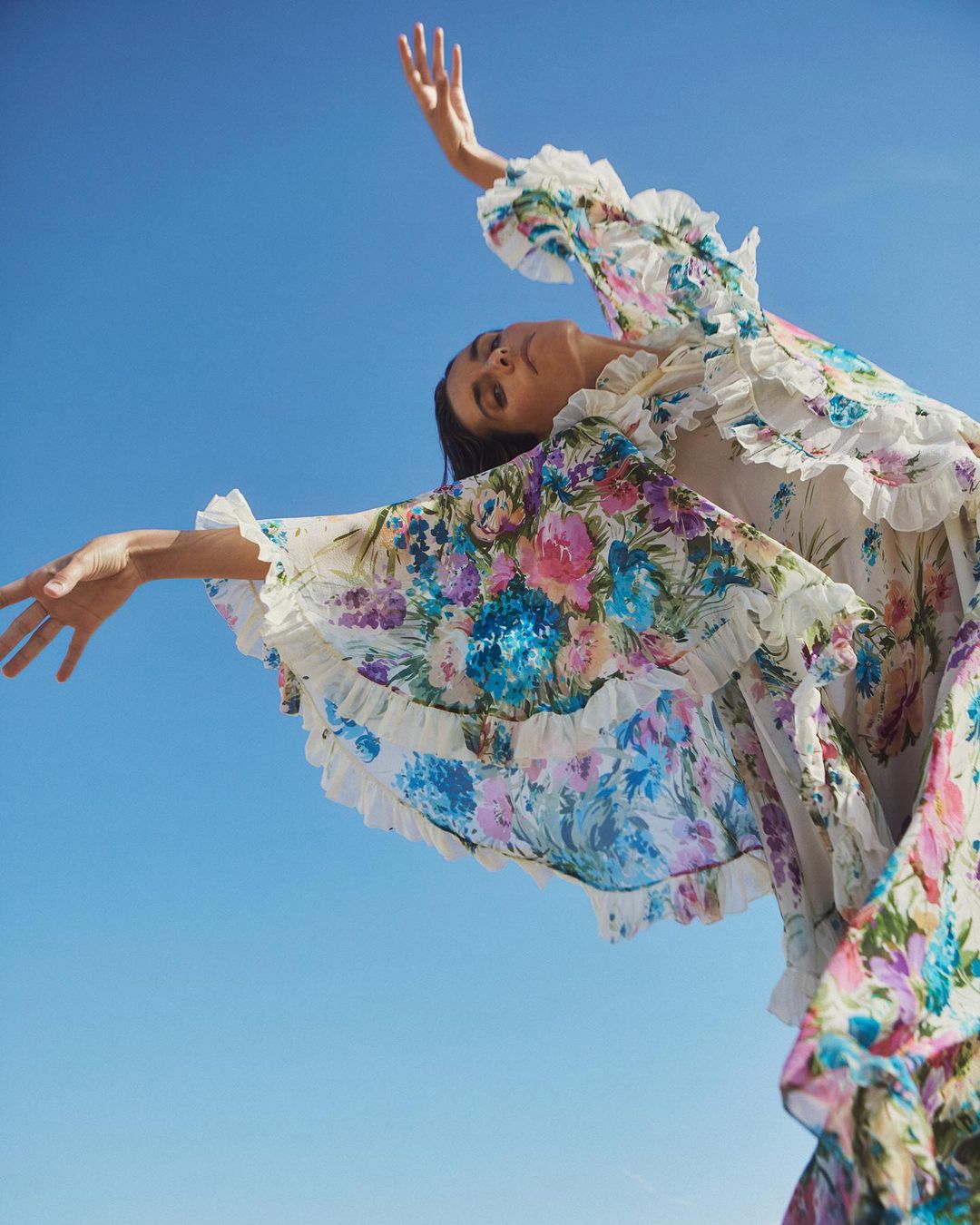 Woman in floral dress with ruffled details poses outdoors against a clear blue sky, arms outstretched, shot from a low angle.