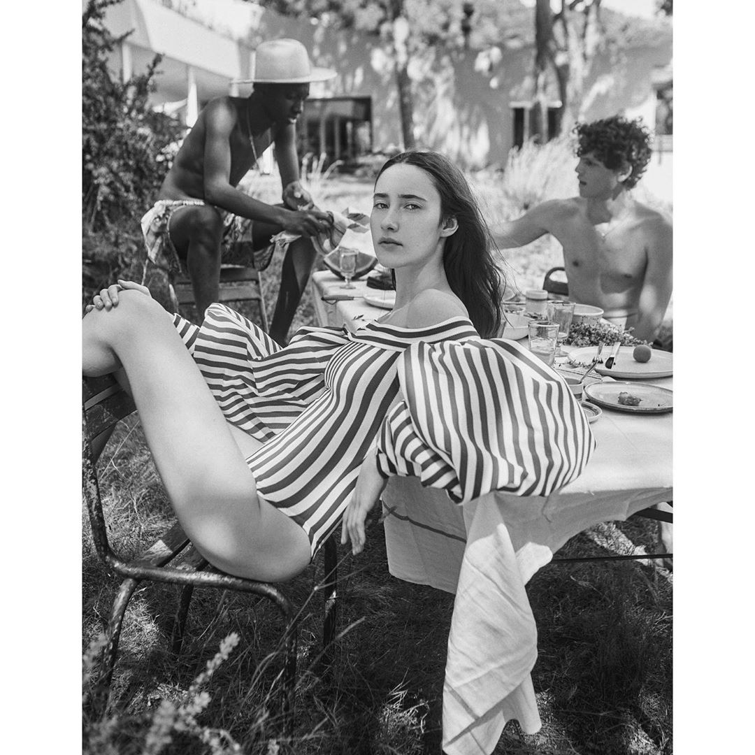 Black and white shot of a woman in a striped bodysuit sitting in a chair, with two shirtless men at a table in the background.