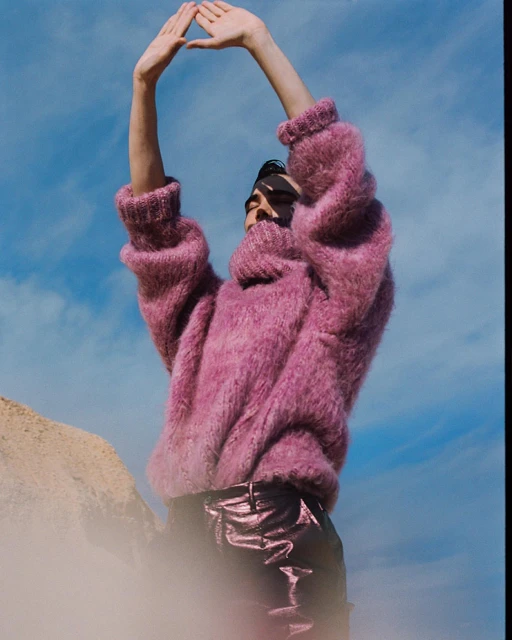A person with arms raised wearing a pink sweater stands on a hill against a blue sky in an outdoor fashion shot.  