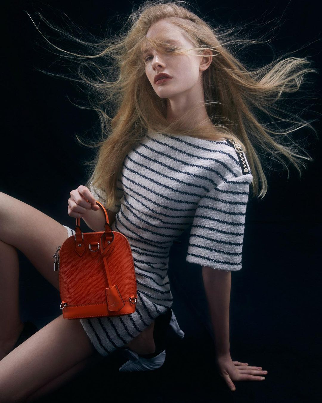 Woman with long blonde hair poses in a striped dress, holding a textured orange handbag against a dark background, fashion studio shot.