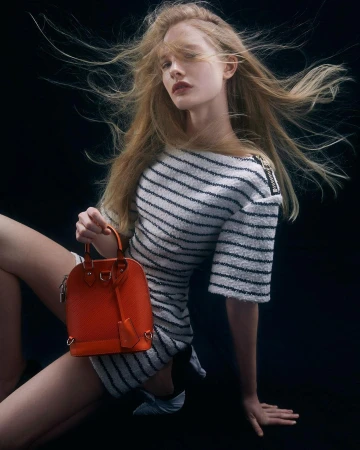 Woman with long blonde hair poses in a striped dress, holding a textured orange handbag against a dark background, fashion studio shot.