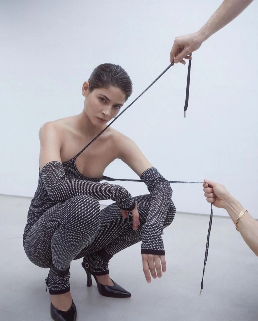 A fair-skinned woman crouches looking at the camera, wearing a patterned catsuit, with hands holding black straps above her.