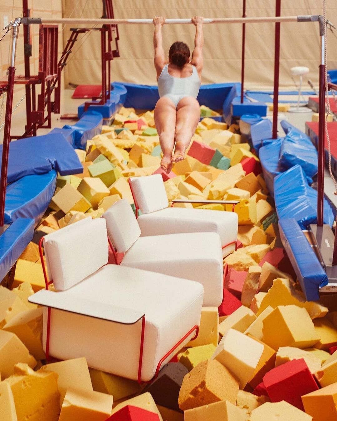 Adult female athlete hangs from a high bar above foam blocks and chairs inside a gym, capturing a unique blend of sport and furniture.