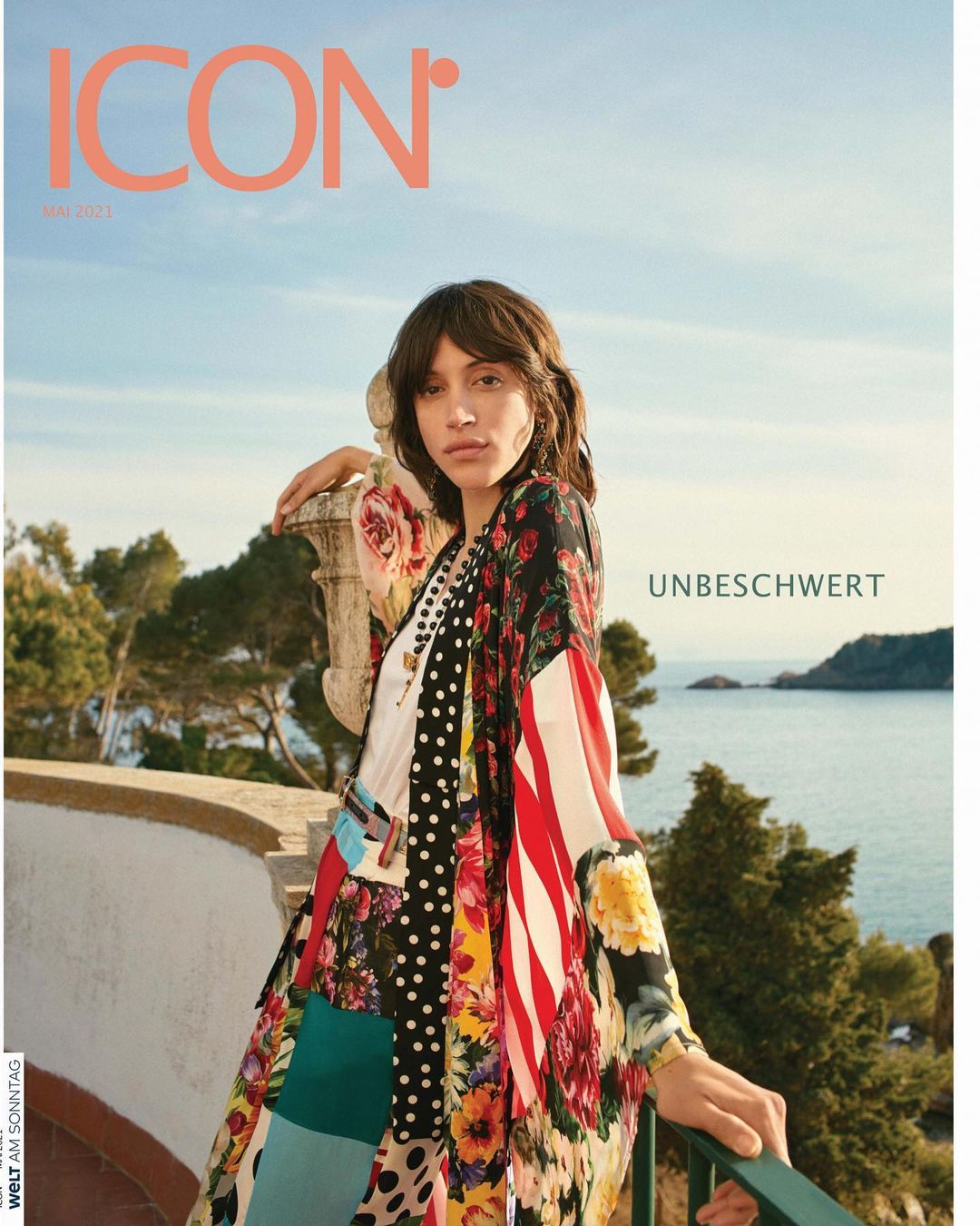 Magazine cover featuring a brunette fashion model in a colorful patterned outfit, posing on a stone balcony with ocean backdrop.