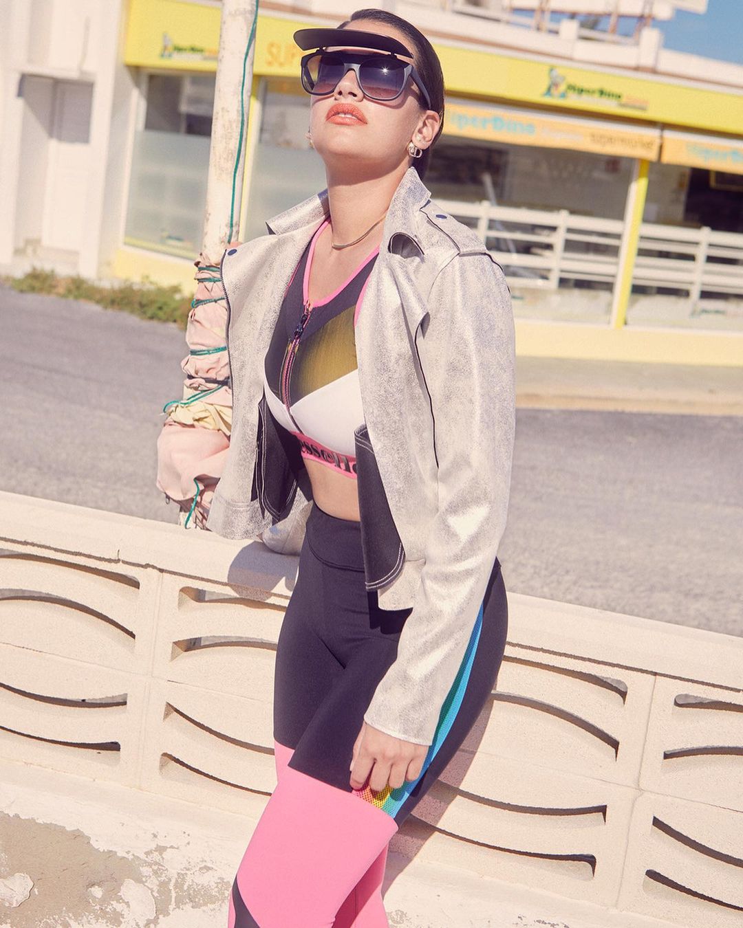 Adult female poses outdoors with a sporty outfit. She wears a silver jacket, sunglasses, and a visor while standing in the sunlight.
