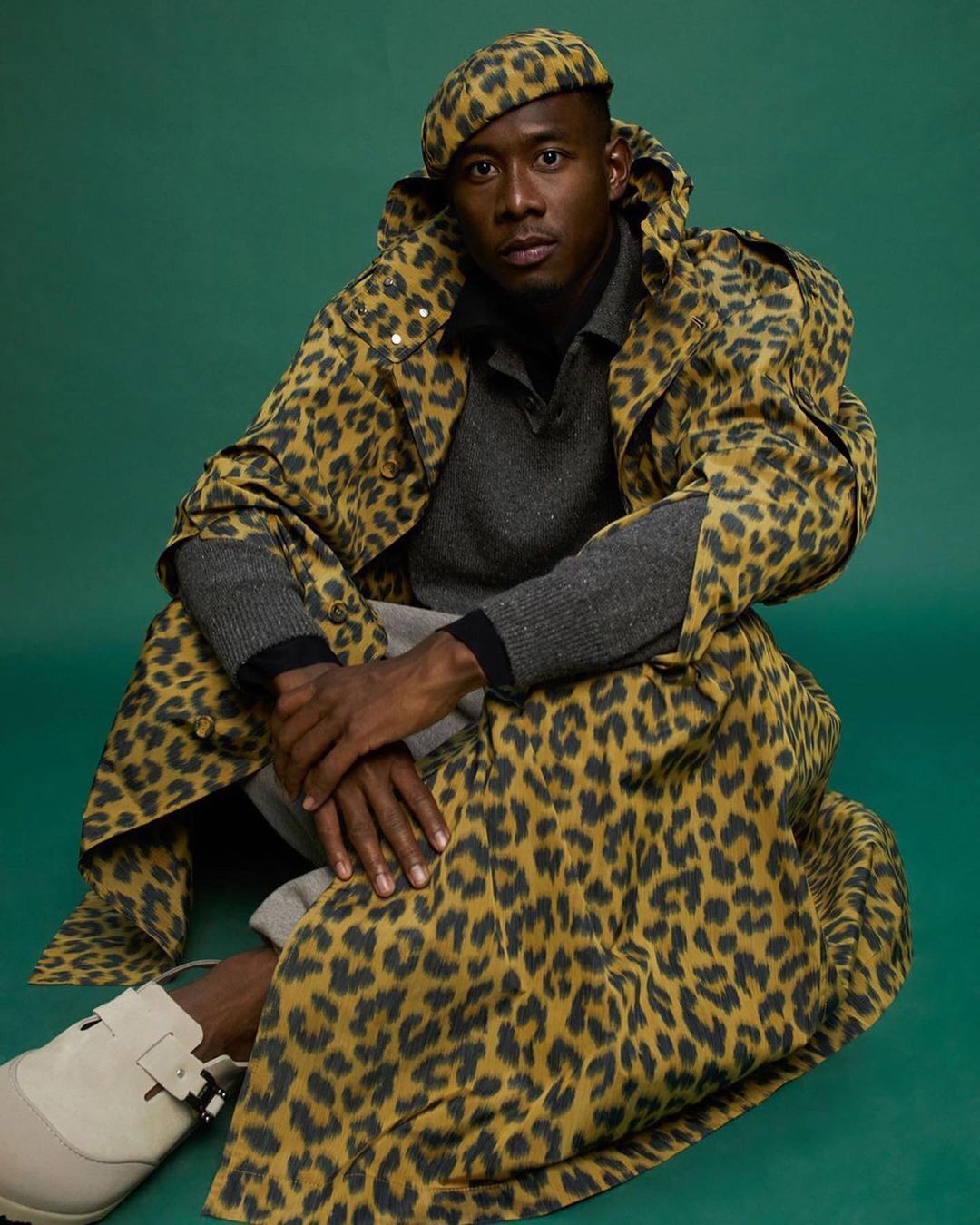 Man in leopard-print beret and coat sitting on a green surface. He wears gray shirt and pants with white shoes and looks at the camera.