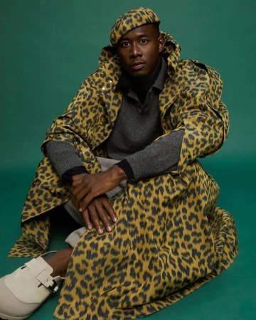 Man in leopard-print beret and coat sitting on a green surface. He wears gray shirt and pants with white shoes and looks at the camera.
