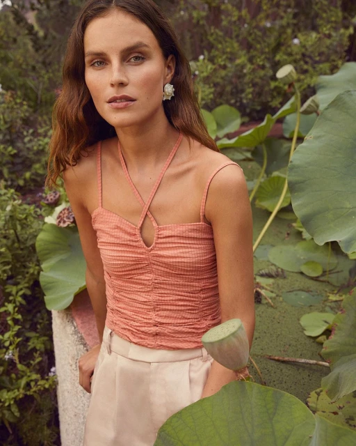 Woman in coral top and cream trousers poses among plants and water lilies in outdoor setting with natural light.