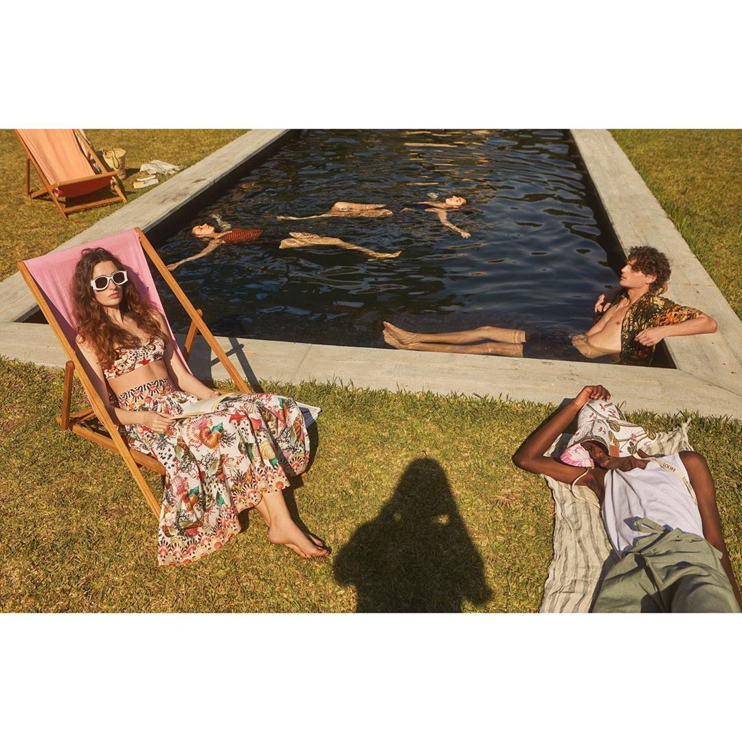 Group of people lounging and swimming at an outdoor pool. A woman in a floral outfit is reclining in a beach chair.
