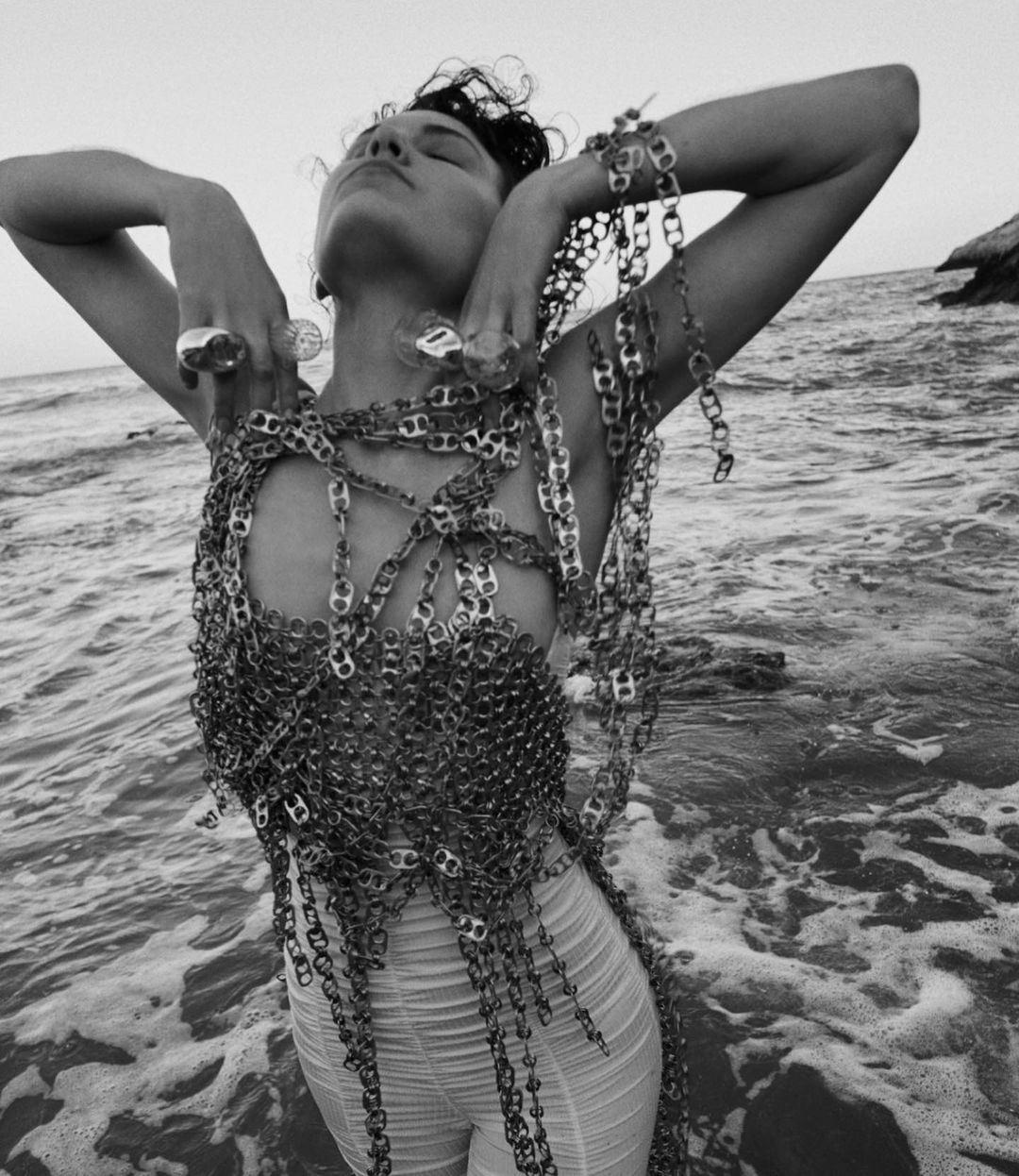A woman in a dress made of aluminum can tabs standing in shallow ocean water with rocks and sky visible in the background in black and white.