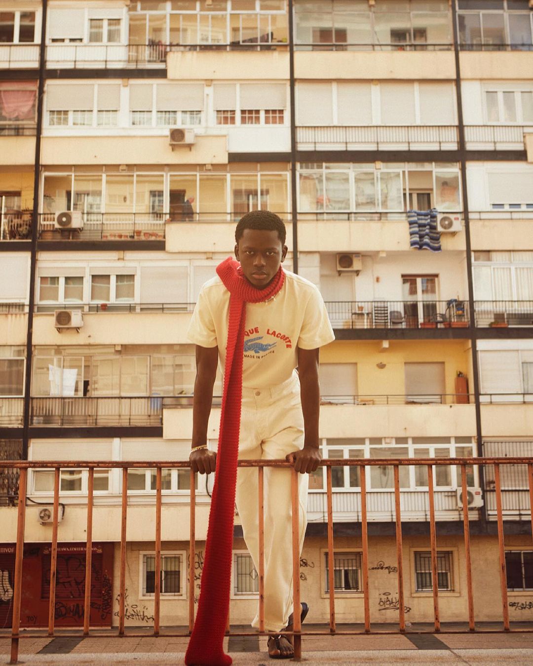 Portrait of Black man leaning on railing in front of a building, wearing a yellow shirt and a long red knit scarf.