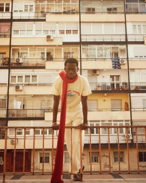 Portrait of Black man leaning on railing in front of a building, wearing a yellow shirt and a long red knit scarf.