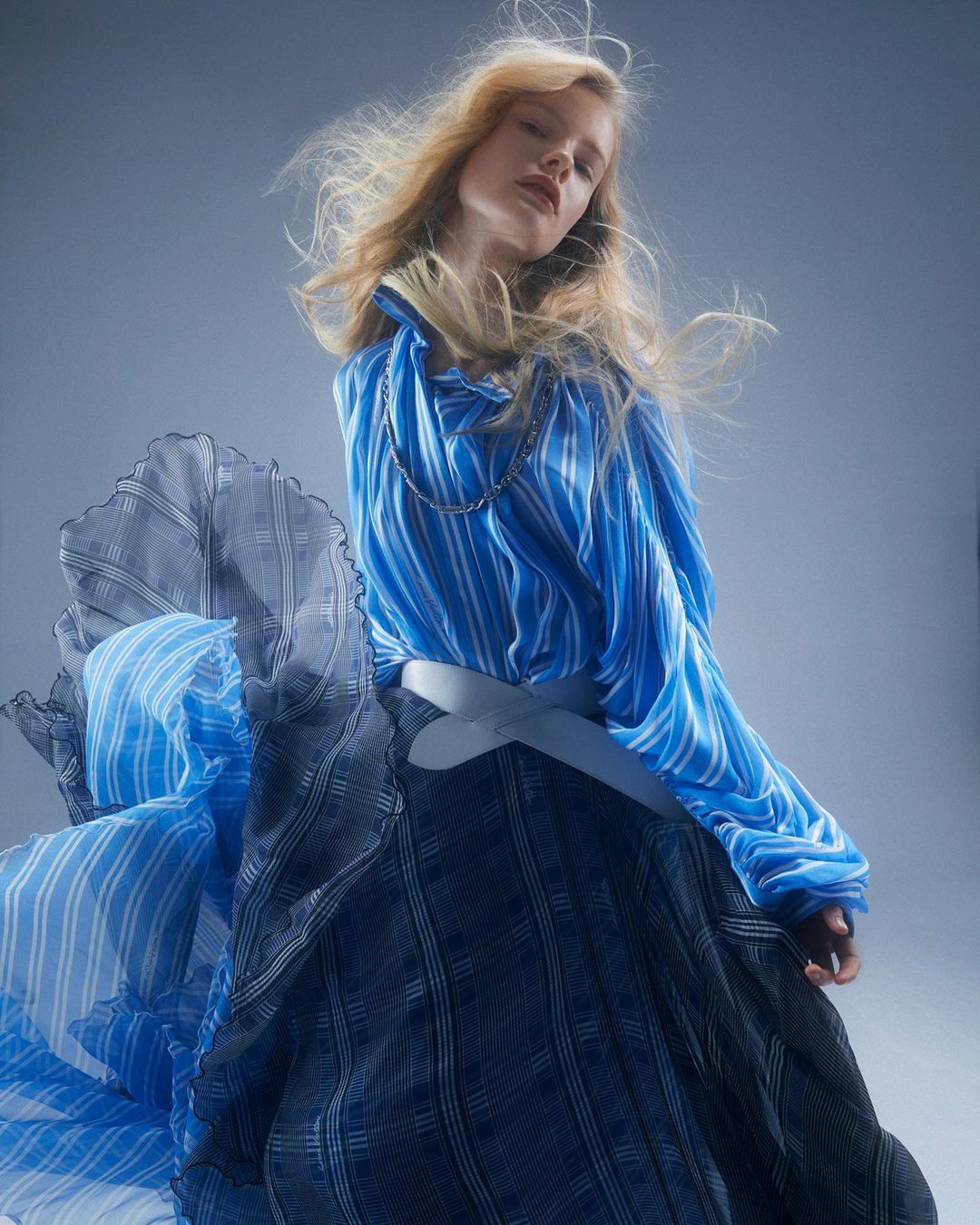 Fashion shot of a blonde model in a blue and black striped outfit with flowing skirt and a neutral backdrop, styled with dynamic posing.