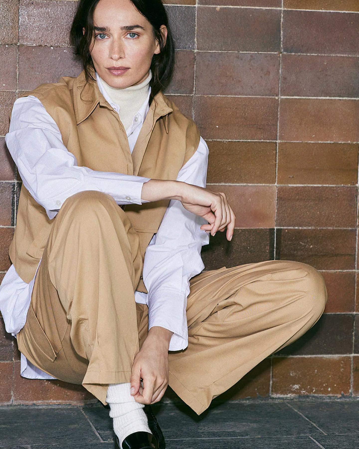A woman in a tan vest and pants poses indoors against a brown-tiled wall, seated with knees up, looking at the camera.