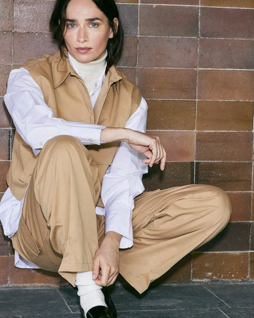 A woman in a tan vest and pants poses indoors against a brown-tiled wall, seated with knees up, looking at the camera.