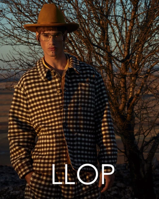 Man in a brown and white checkered coat and hat stands in front of a bare tree during golden hour with a tan field in the background.