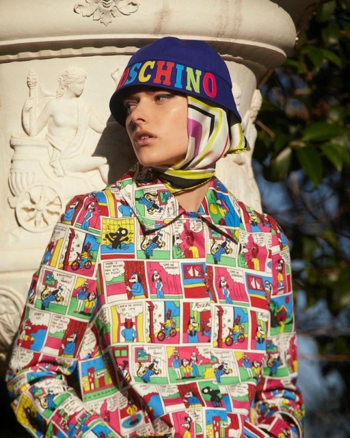Woman poses outdoors wearing a colorful, comic-style jacket and blue bucket hat with multicolored Moschino lettering in an outdoor environment.